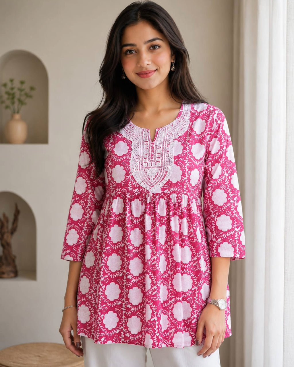 A woman wearing a pink and white floral print tunic top with white embroidery around the neckline, standing in a brightly lit indoor setting.