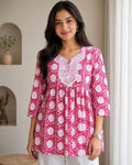 A woman wearing a pink and white floral print tunic top with white embroidery around the neckline, standing in a brightly lit indoor setting.