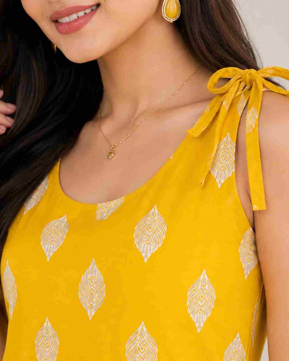 A close-up of a woman wearing a bright yellow sleeveless top with a repeating white leaf-like pattern and tie-shoulder straps, along with gold jewelry.