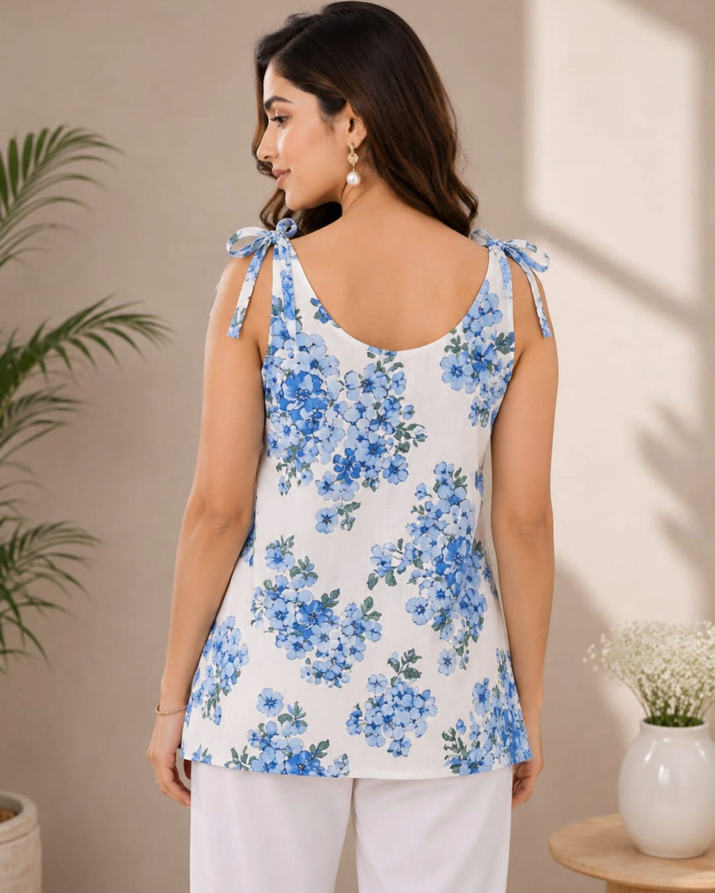 A woman shown from the back wearing a sleeveless white Kurti with a blue floral print and tie-shoulder straps, paired with white pants in a softly lit indoor setting.