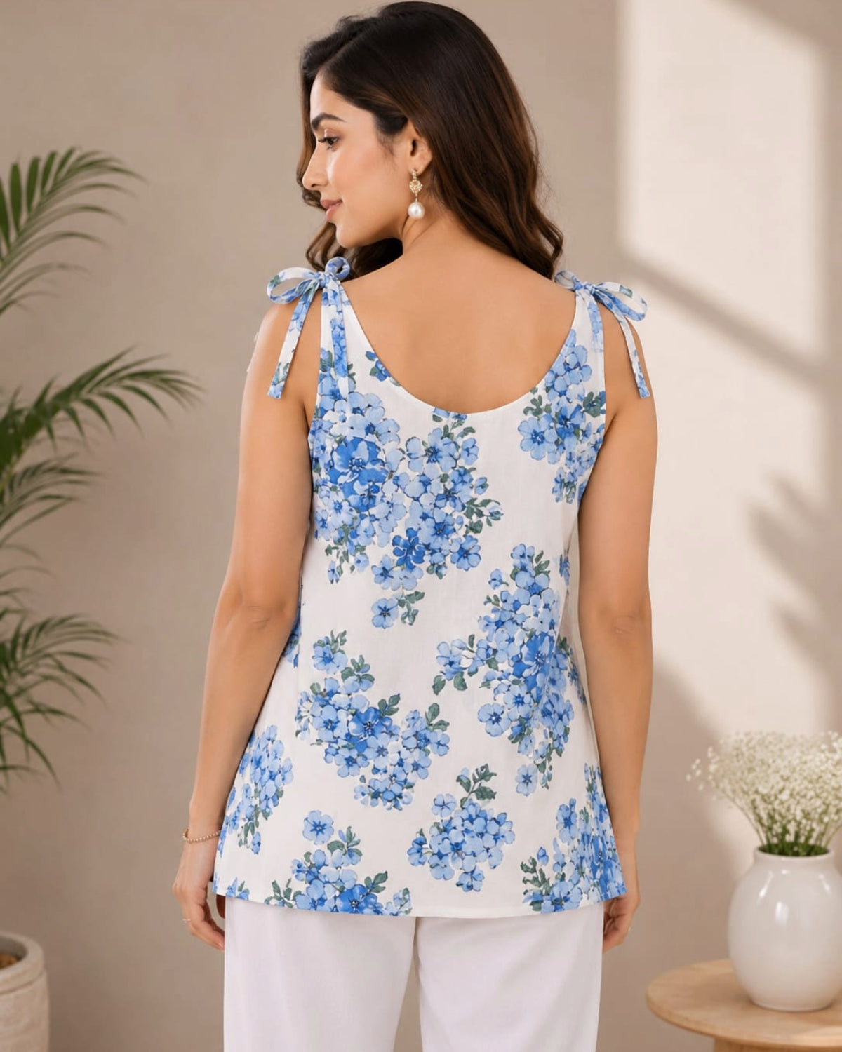 A woman shown from the back wearing a sleeveless white Kurti with a blue floral print and tie-shoulder straps, paired with white pants in a softly lit indoor setting.