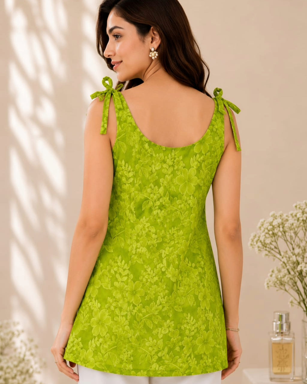 A woman wearing a bright lime green floral lace sleeveless top, shown from the back. The top features tie-shoulder straps and a scoop neckline, photographed in a softly lit indoor setting with decorative flowers in the background.