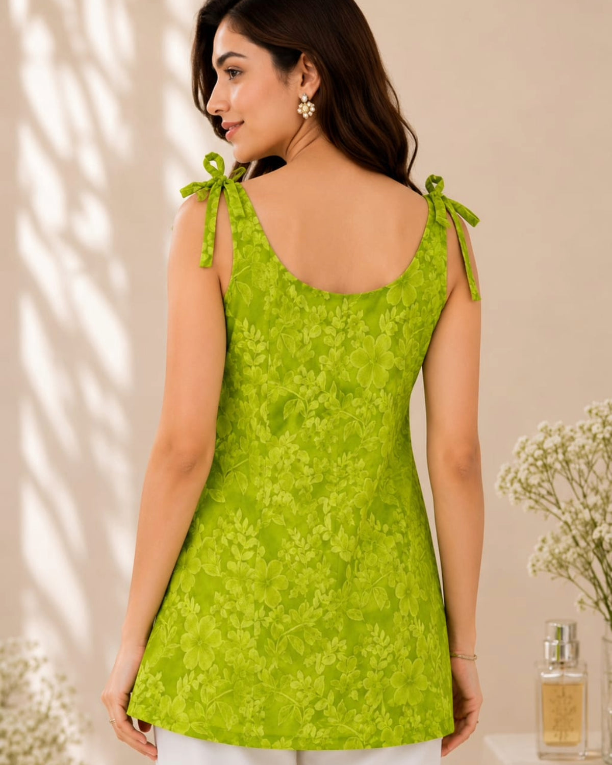 A woman wearing a bright lime green floral lace sleeveless top, shown from the back. The top features tie-shoulder straps and a scoop neckline, photographed in a softly lit indoor setting with decorative flowers in the background.