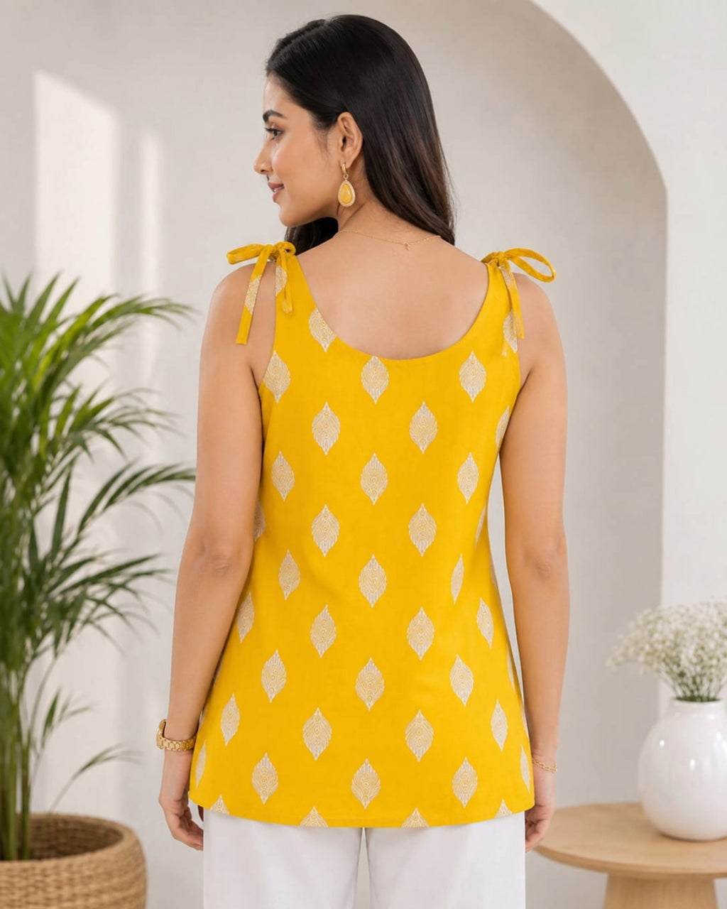 Elegant female model wearing a mustard yellow sleeveless cotton top with white geometric diamond print, tie-shoulder straps, paired with white pants, modern indoor lifestyle background, front view.