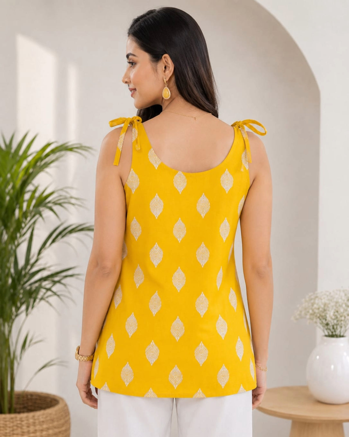 Elegant female model wearing a mustard yellow sleeveless cotton top with white geometric diamond print, tie-shoulder straps, paired with white pants, modern indoor lifestyle background, front view.