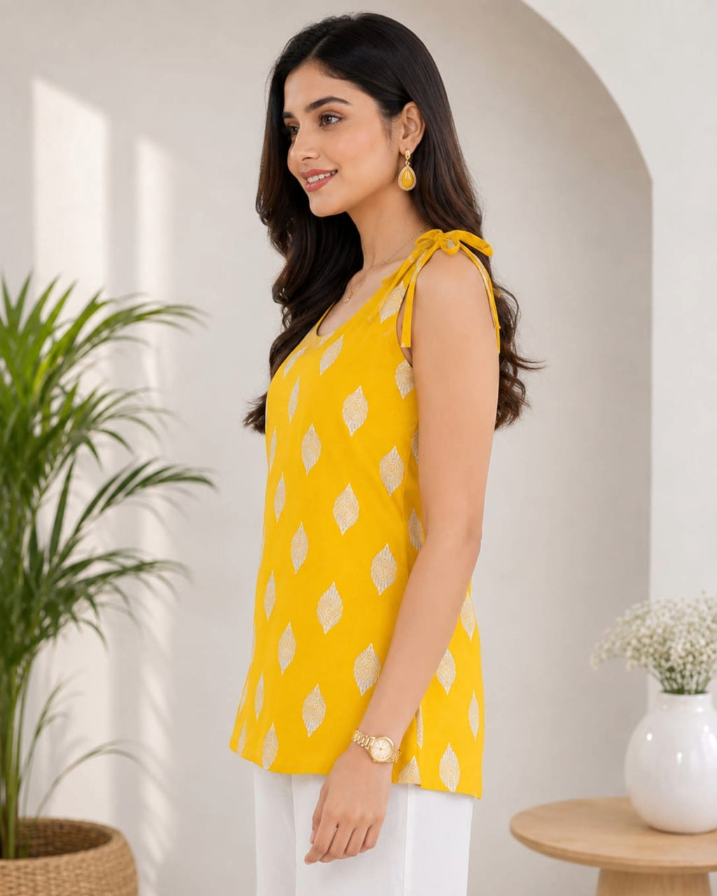 Elegant female model wearing a mustard yellow sleeveless cotton top with white geometric diamond print, tie-shoulder straps, paired with white pants, modern indoor lifestyle background, front view.